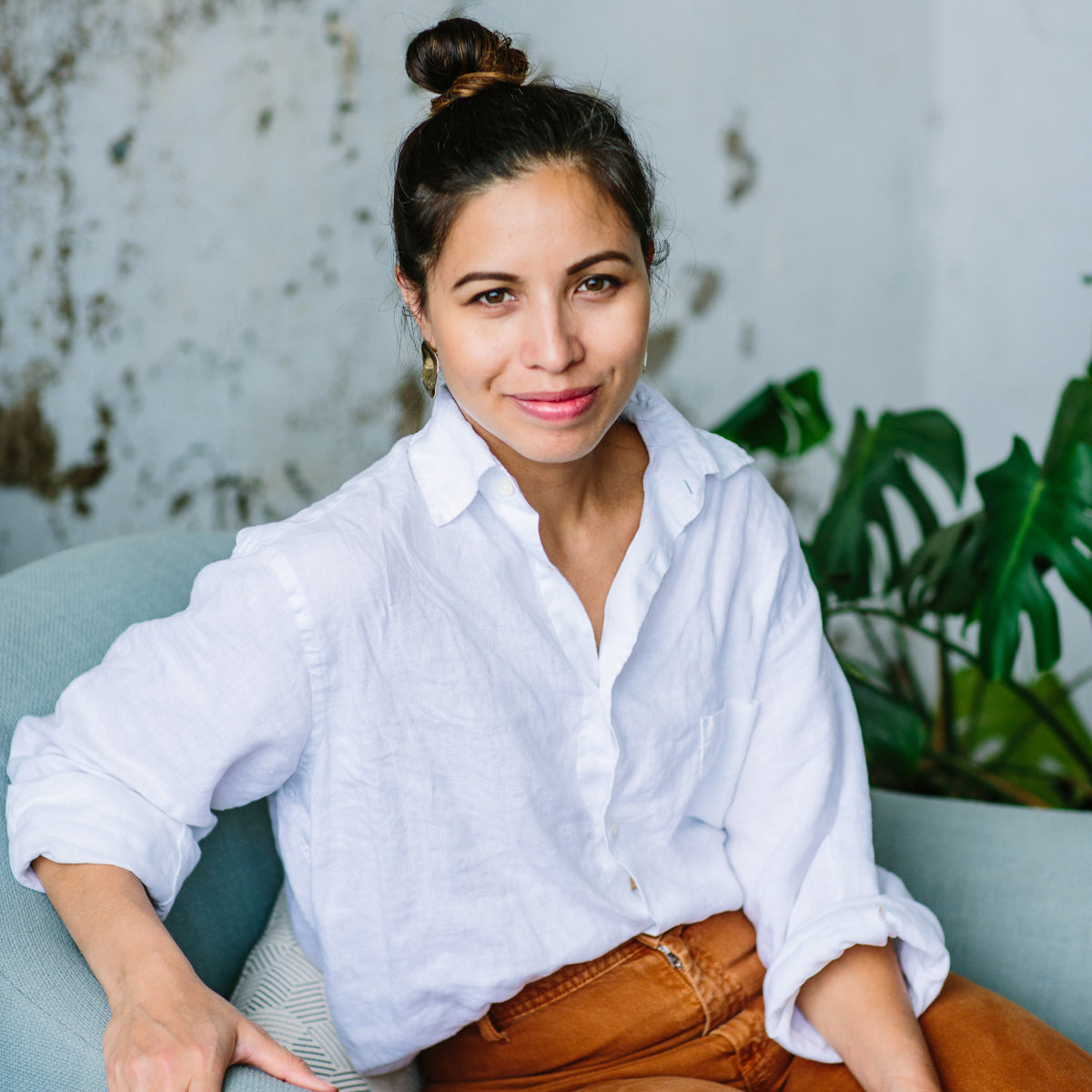 A woman in a white shirt and brown pants sitting on a light blue chair, smiling with a plant in the background.