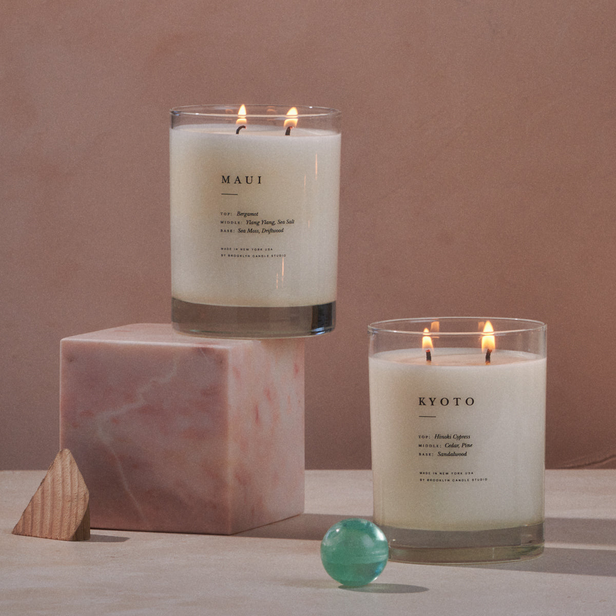 Two lit candles labeled Maui and Kyoto placed on a surface with a pink stone cube and a green marble.