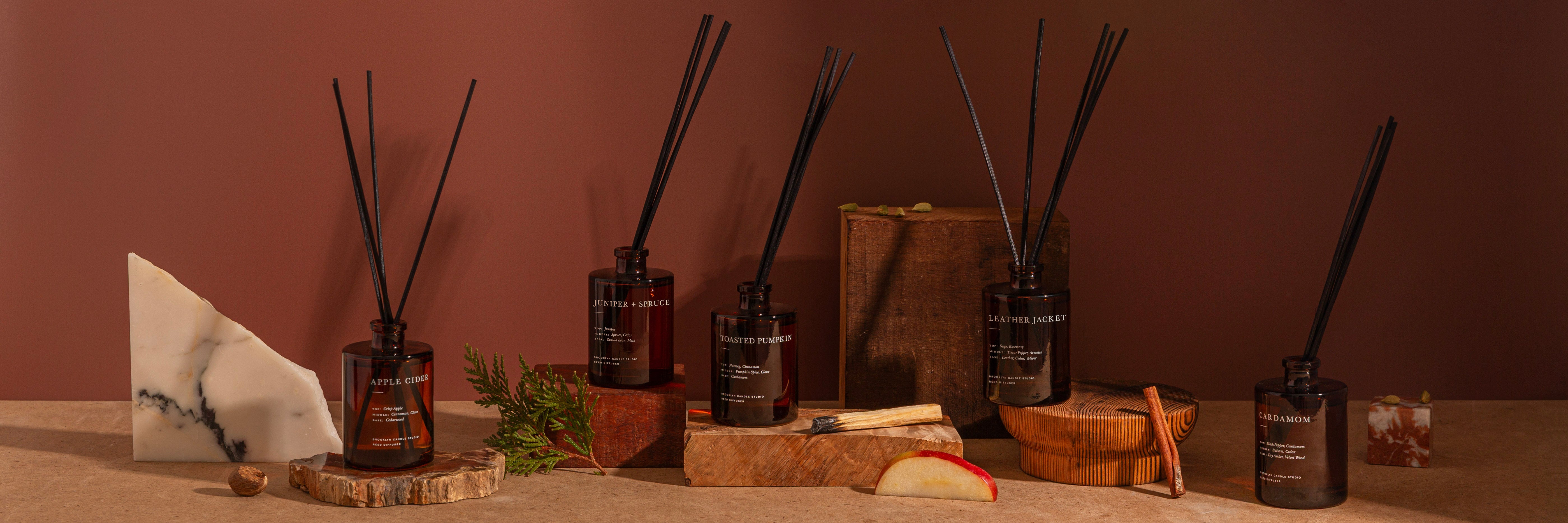 A styled product display of several amber glass reed diffusers with black reeds, arranged on wooden blocks against a warm terracotta background. Decorative props include a white marble slice, a small green sprig, a slice of apple, cinnamon sticks, and a brown box.